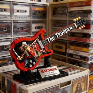 Puede incluir: Un coleccionable en forma de guitarra roja y negra con el logotipo de Iron Maiden y las palabras "The Trooper". La guitarra tiene un soporte negro y una placa conmemorativa que dice "Iron Maiden 1975-2025 50th Anniversary."