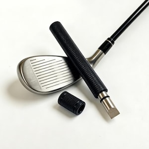 以下が含まれることがあります： 黒いテクスチャードグリップとシルバーのヘッドを備えたゴルフクラブ。別の黒いテクスチャードグリップも存在します。クラブは白い表面に配置され、そのデザインと特徴を際立たせています。