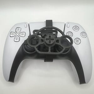 Puede incluir: Un accesorio de volante negro impreso en 3D montado en un mando de videojuegos blanco y negro. El accesorio presenta un marco circular con un diseño de cruz central, asegurado al mando mediante una correa negra que cruza el centro.