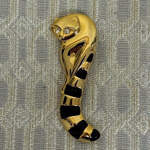 May include: A gold-coloured raccoon brooch with a sleek, curved design. The raccoon's eyes are accented with small, sparkling rhinestones. The tail features alternating black and gold stripes, adding a touch of contrast to the piece. A unique and eye-catching accessory.