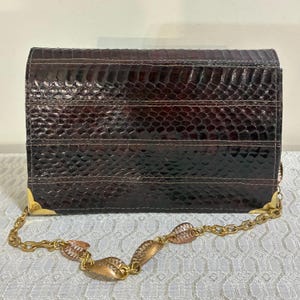 May include: A rectangular dark brown snakeskin clutch bag featuring a textured scale pattern. The bag includes gold-tone metal corner protectors and a decorative gold-tone chain strap composed of interconnected leaf-shaped metal links.