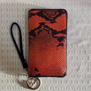 Puede incluir: Una cartera rectangular con cremallera y un exterior con estampado de piel de serpiente en naranja y negro. La cartera incluye una correa de muñeca negra unida a un dije circular de metal plateado con texto grabado para facilitar su transporte.