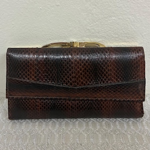 May include: A brown and black snakeskin wallet with a gold-coloured clasp. The wallet has a rectangular shape with a fold-over flap. The snakeskin pattern is visible across the entire surface of the wallet.