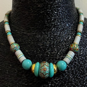 May include: A necklace featuring a sequence of cylindrical light grey beads, round turquoise beads, and two spherical beads decorated with a colourful floral pattern. The beads are separated by small gold-toned metal spacers and rings.