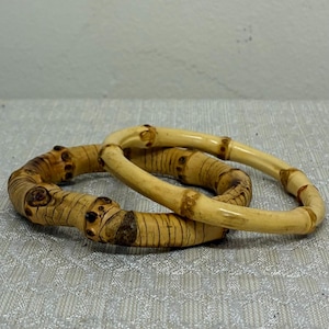 Op de afbeelding: Twee bamboe armbanden. Eén armband is lichtbruin van kleur met donkerbruine knopen en lijnen. De andere armband is lichtbruin van kleur. Beide armbanden zijn rond en hebben een natuurlijke, organische uitstraling.