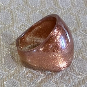 May include: A thick, rounded ring with a smooth, glossy finish. The band features a translucent, warm copper-toned base embedded with fine, reflective glitter particles that catch the light, creating a shimmering, textured appearance throughout.
