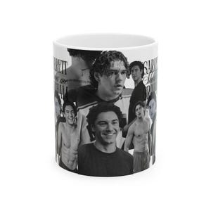 Può includere: Una tazza in ceramica bianca con un collage di foto in bianco e nero di una persona con capelli ricci. Il design include diversi ritratti e il testo ripetuto "GARRETT Show me GRAHAM" in un carattere serif stampato sulla superficie esterna.