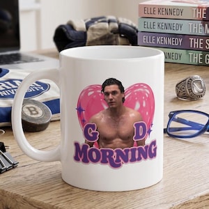 May include: A white ceramic mug featuring a graphic of a shirtless man centered in front of a large pink heart. The text "G D MORNING" is printed in purple letters below the heart. The design includes small purple star accents on the sides.