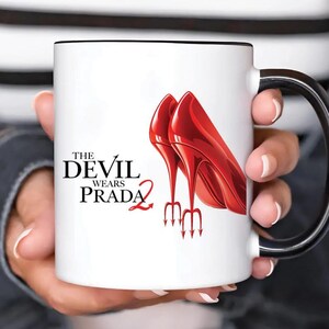 Op de afbeelding: Witte keramische mok met een zwarte handgreep en rand. De mok heeft de tekst "The Devil Wears Prada 2" en een illustratie van twee rode hoge hakken met drietandhakken.