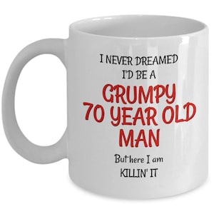 May include: White ceramic mug with a curved handle. The mug features the text "I NEVER DREAMED I'D BE A GRUMPY 70 YEAR OLD MAN But here I am KILLIN' IT" in black and red lettering. A humorous gift.