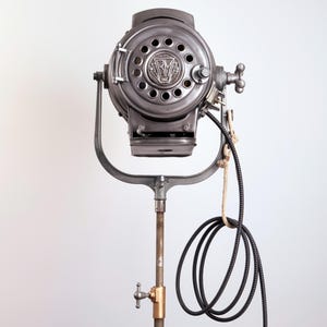 Könnte beinhalten: Vintage Industrie-Scheinwerfer mit rundem Kopf und den Buchstaben "R M". Der Scheinwerfer ist auf einem Metallständer mit Messingarmaturen und einem aufgerollten schwarzen Netzkabel montiert. Höhe ca. 150 cm.