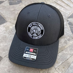 May include: Black trucker hat with a white embroidered logo that says "Old Hollywood Light Company". The hat has a black mesh back and a black adjustable strap with a red "R" and the text "Richardson 112 Original Trucker Adjustable".