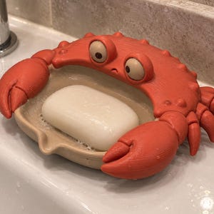 May include: A bright orange crab-shaped soap dish with two large eyes and textured claws. The dish features a beige interior basin holding a single, smooth, white rectangular bar of soap. The crab is positioned with its claws extended forward.
