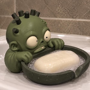 May include: An olive green zombie-themed soap dish featuring a cartoonish character with large white eyes, small black hair spikes, and a stitched forehead, holding a dark green oval tray containing a single white bar of soap with bubbles.