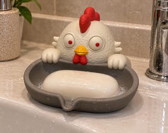 Könnte beinhalten: Eine graue Seifenschale in Form eines Huhns mit großen Augen, einem roten Kamm und einem gelben Schnabel. Die Hühnerfigur hält das ovale graue Becken mit ihren weißen Flügeln. Ein glattes, weißes Stück Seife liegt mit Wassertropfen darin.