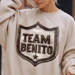 May include: Beige sweatshirt featuring a shield-shaped patch with the text "TEAM BENITO". The patch has a brown border and a star. The sweatshirt is a neutral colour and has long sleeves.