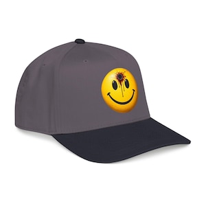 Puede incluir: Gorra de béisbol gris y azul marino con un diseño de cara sonriente amarilla con un agujero de bala. La cara sonriente tiene un contorno negro y un efecto de sangre. La gorra tiene una visera curva.