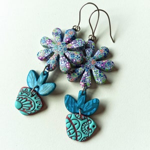 May include: A pair of dangle earrings featuring a floral design. The top part of the earrings is a large, multi-coloured flower with a blue, green and purple pattern. The bottom part of the earrings is a smaller, blue and green flower with a red and white pattern.