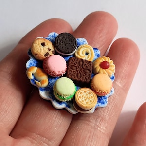 May include: Miniature assortment of cookies and pastries on a blue and white plate. Includes a chocolate chip cookie, an Oreo, a pink macaron, and a croissant. The plate is held in a person's hand.