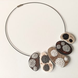 Puede incluir: Un collar negro con un cierre plateado presenta cinco colgantes de cerámica marrón, blanco y negro. Cada colgante tiene forma ovalada y presenta un diseño único de círculos blancos y negros.
