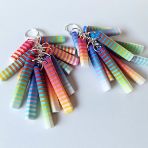 Puede incluir: Un par de pendientes colgantes de rayas de colores. Cada pendiente tiene múltiples barras delgadas de arcilla polimérica rayadas en tonos de rojo, naranja, amarillo, verde, azul, morado y rosa. Las barras están unidas a un gancho plateado.