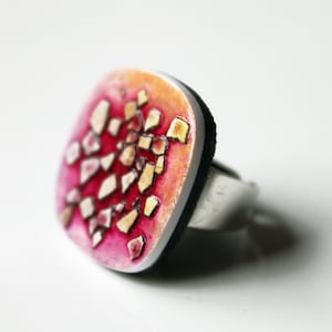 Puede incluir: Un anillo cuadrado con un diseño de mosaico rosa, naranja y blanco. El anillo está hecho de metal y tiene una banda negra.