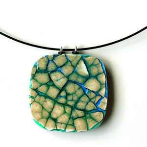 Può includere: Una collana con un ciondolo quadrato in ceramica craquelé in tonalità di verde, blu e bianco. Il ciondolo è attaccato a un cordoncino nero.