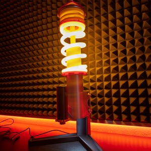 Könnte beinhalten: Eine einzigartige Schreibtischlampe im Design einer Auto-Stoßdämpferfeder. Sie hat eine weiße, spiralförmige Lichtquelle in einem roten und gelben Gehäuse, montiert auf einem schwarzen Sockel. Die Lampe ist angeschlossen und leuchtet.