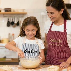 May include: Two aprons featuring vertical pinstripes. The larger maroon apron has the embroidered text Sarah Cookie Queen. The smaller blue and white striped apron has the embroidered name Sophia. Both aprons have adjustable neck straps and waist ties.