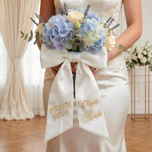 Puede incluir: Un ramo de novia con hortensias azules, rosas blancas y lavanda, atado con un gran lazo de satén blanco. El lazo tiene bordado dorado: "PROM 2026" y "Olivia & Noah". La persona lleva un vestido blanco.