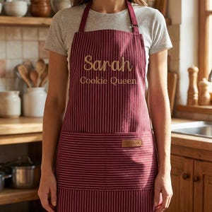 May include: A burgundy apron with thin white vertical stripes, featuring the embroidered text Sarah and Cookie Queen in gold thread. The apron includes a large front pocket with a small rectangular brown leather-like brand tag attached.