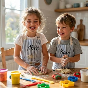 May include: Two children wearing matching blue and white pinstriped aprons with the names Alice and Sam embroidered in black script. They are standing at a wooden table covered in colorful modeling clay, plastic tools, and cookie cutters.