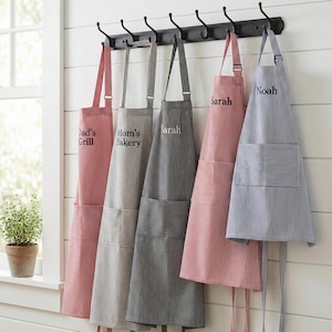 May include: Five striped aprons in red, gray, charcoal, and blue hanging on a black wall-mounted rack with hooks. Each apron features a front pocket and embroidered text including Dad's Grill, Mom's Bakery, Sarah, and Noah in various fonts.