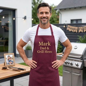 May include: A burgundy apron with thin white vertical pinstripes featuring the embroidered gold text Mark Dad & Grill Hero. The apron has a neck strap and a large front pocket, designed for cooking and grilling activities in an outdoor setting.