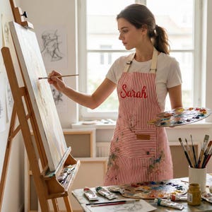 May include: A red and white striped apron featuring the name Sarah embroidered in red script on the chest. The apron includes a front pocket and adjustable straps, worn over a white t-shirt while painting on a wooden easel with a palette.