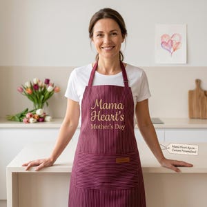 May include: A burgundy apron with thin white vertical stripes, featuring the gold text "Mama Heart's Mother's Day" on the chest and a front pocket with a small rectangular brown leather-like patch. The apron has adjustable neck and waist straps.