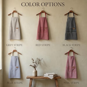 May include: Five aprons hanging on wooden hangers, labeled Color Options. The aprons feature vertical stripes in grey, red, black, blue, and pink. Each apron has a front pocket with horizontal stripes and a small rectangular patch on the pocket.