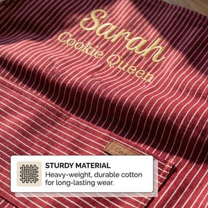May include: A red and white pinstriped apron featuring gold embroidered text that reads Sarah Cookie Queen. The apron includes a front pocket with a small brown rectangular tag. A white informational box is overlaid with the text STURDY MATERIAL.
