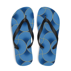 Op de afbeelding: Een paar slippers met een herhalend patroon van blauwe concentrische cirkels en dunne gouden gebogen lijnen. Het schoeisel heeft zwarte rubberen teenslippers bevestigd aan de zolen, die een verloop van licht- naar donkerblauwe tinten vertonen.