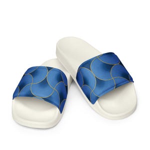 Op de afbeelding: Een paar witte slippers met een brede band met een blauw en goud cirkelvormig golfpatroon. Het ontwerp bestaat uit overlappende blauwe cirkels met fijne concentrische lijnen en dunne gouden omlijningen op een donkerblauwe achtergrond.