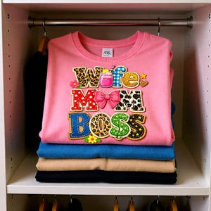 Op de afbeelding: Een roze crewneck t-shirt met de woorden "Wife Mom Boss" in een kleurrijk, decoratief ontwerp. Het shirt is op andere opgevouwen shirts in een kast gestapeld.