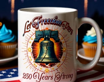 May include: White ceramic mug featuring the Liberty Bell with the text "Let Freedom Ring" and "250 Years Strong." The design includes a burst of light and stars. The mug is on a red, white, and blue napkin with cupcakes in the background.