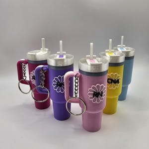 May include: A collection of colorful tumbler keychains in shades of pink, purple, yellow, and blue. Each tumbler features a white daisy design with the letters "RN" or "DNA" and a silver keychain. The tumblers have white lids and straws.