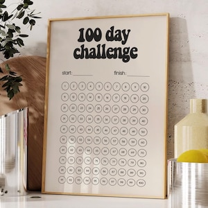 May include: A framed print with the text "100 day challenge" in a bold, retro font. The print has a grid of 100 circles for tracking progress. The frame is gold, and the background is a light beige color.