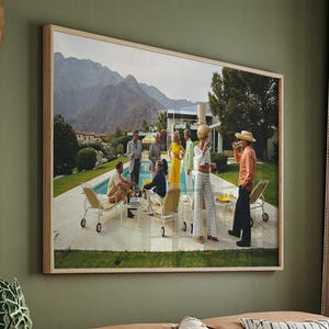 May include: A framed print depicts a retro outdoor gathering by a pool, with people in vintage attire. The scene is set against a backdrop of mountains and a mid-century modern home. The print hangs above a brown leather sectional sofa.