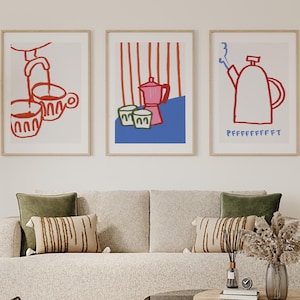 May include: Three framed art prints with coffee-themed illustrations in red and white. One print shows a coffee maker pouring into cups, another features a pink coffee pot and cups, and the third depicts a teapot with steam and the text "PFFFFFFFFFT".