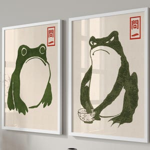 May include: Two framed art prints featuring green frog illustrations. The frogs are depicted in a minimalist style with a Japanese aesthetic. Each print has a white frame and a red Japanese character in the upper right corner.