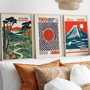 Könnte beinhalten: Drei gerahmte Kunstdrucke mit japanisch inspirierten Designs. Die Drucke zeigen Illustrationen eines japanischen Gartens, einer roten Sonne mit schwarzen Punkten und dem Berg Fuji. Die Farbpalette umfasst Orange-, Grün- und Blautöne. Die Drucke werden über einem Bett mit Kissen angezeigt.