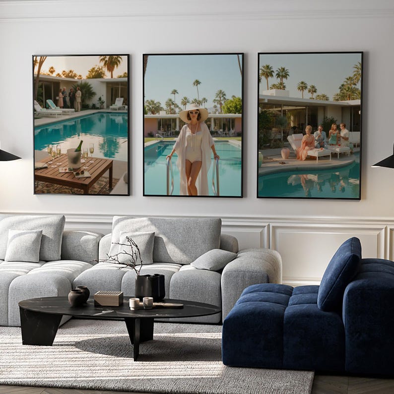 May include: Set of three framed art prints depicting scenes by a swimming pool. One shows a woman in a white outfit, another a table with champagne, and the third a group of people. The prints have black frames and are displayed above a gray sofa.