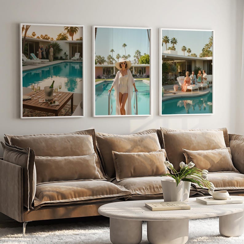 May include: Three framed art prints depicting a retro poolside scene. The prints show a pool, people, and a person in a white outfit. The prints are above a brown sofa with pillows and a white coffee table.